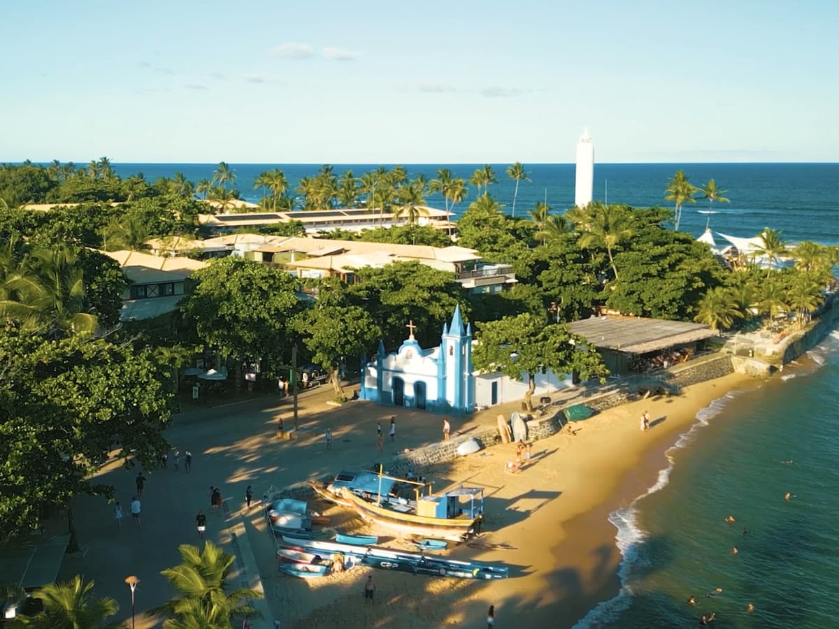 Vista aérea da vila de Praia do Forte no fim da tarde: a igreja de São Francisco de Assis, barcos na areia, o casario entre os coqueiros e o farol junto ao mar